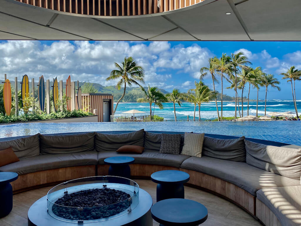 Interior of hotel open to a view of turquoise ocean, surfboards and palm trees blowing in the wind