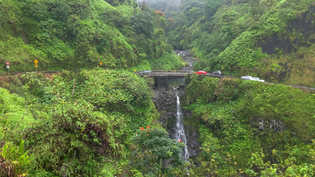 traffic slows at a narrow bridge above a waterfall on maui's famous road to hana