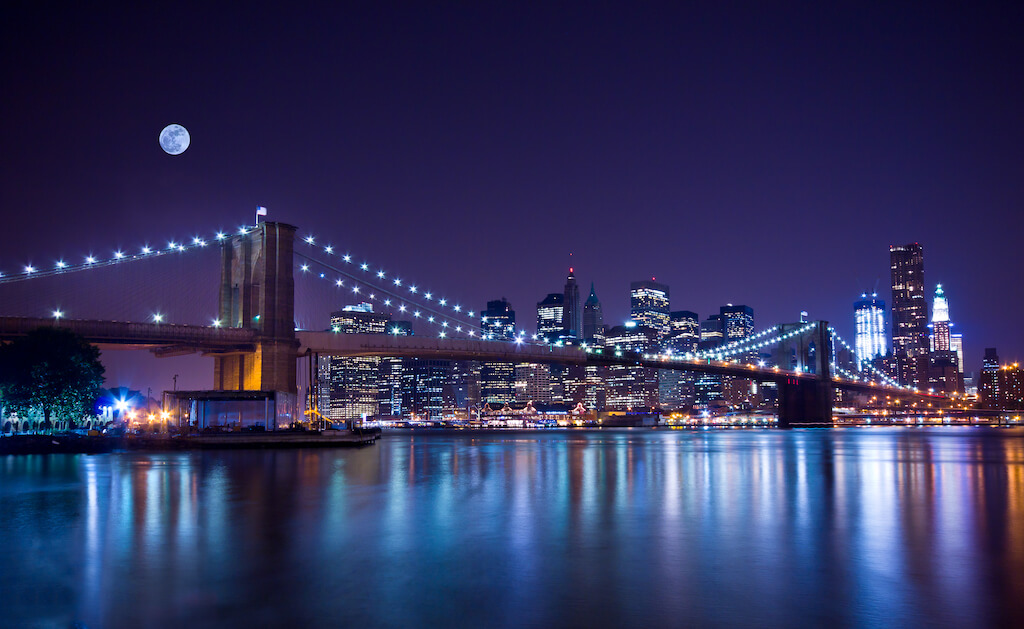 Brooklyn Bridge lit up at night with full moon and cityscape in background